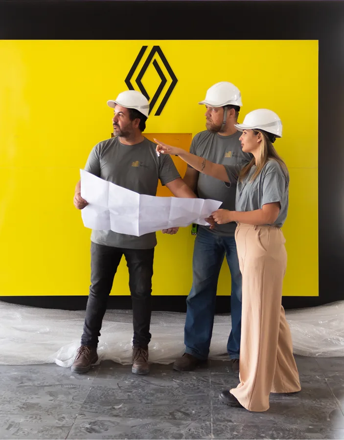 Engenheiros analisando planta de obra em frente a painel amarelo com logotipo