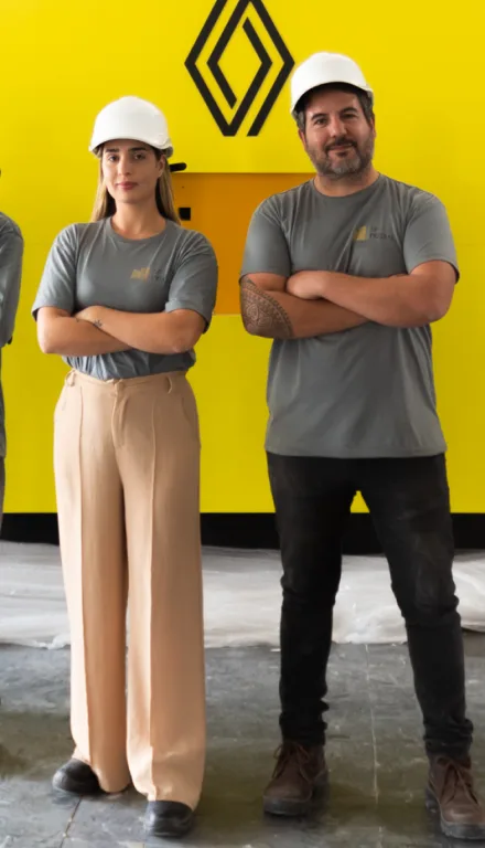Homem e mulher com capacetes de segurança posando em frente a parede amarela com logotipo